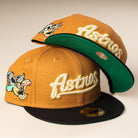 Two El Gallo Canvas Bronze Black Houston Astros 5950 Fitted New Era Hats: both mustard-yellow with black brims, green underbrims, and embroidered logos; one is upright showing the front, the other tilted to show the underside and side patch.