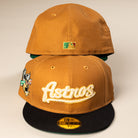 Two El Gallo Canvas Bronze Black Houston Astros New Era Hat 5950 Fitted caps are shown; the front cap features "Astros" in gold, a cartoon patch on the side, and an MLB sticker on the brim. The back cap displays the Houston Astros MLB logo.