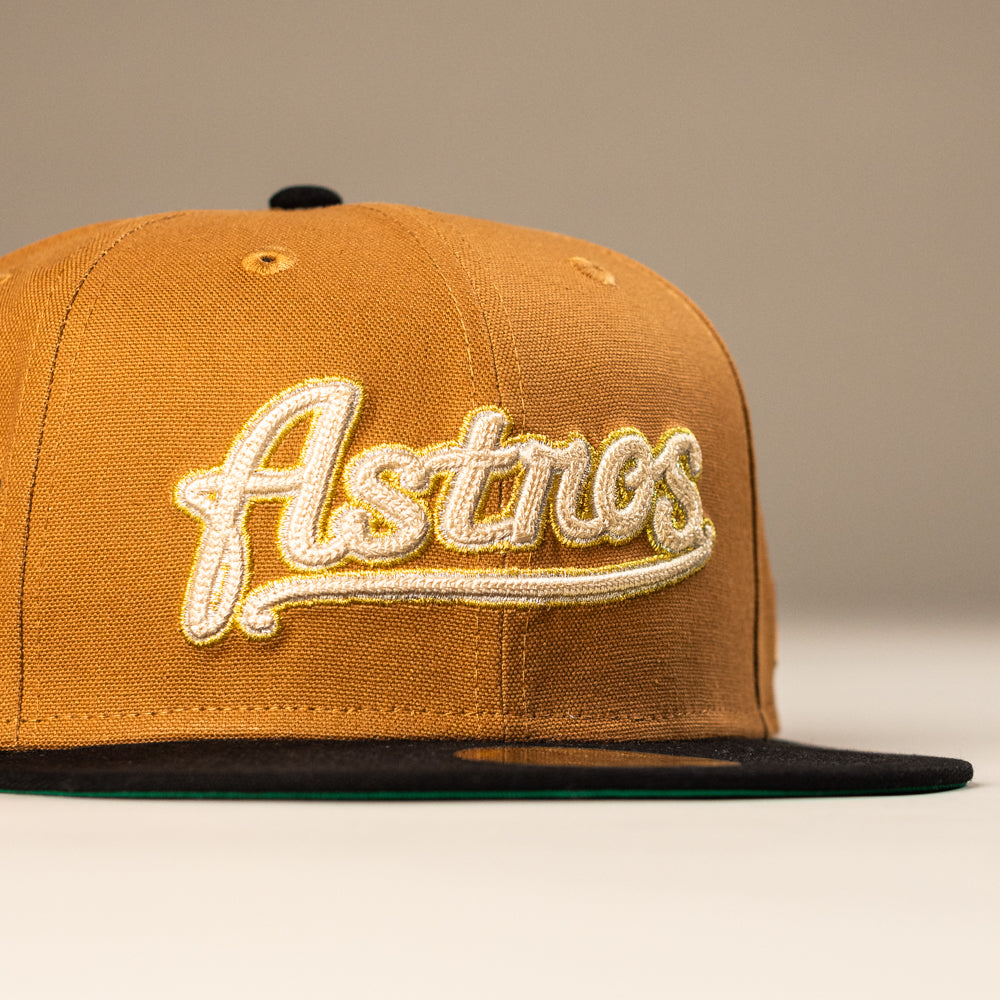 The El Gallo Canvas Bronze Black Houston Astros New Era 5950 Fitted Hat features bronze and black colors, with "Astros" embroidered in white and gold script on the front, a flat brim, and is shown against a neutral background.