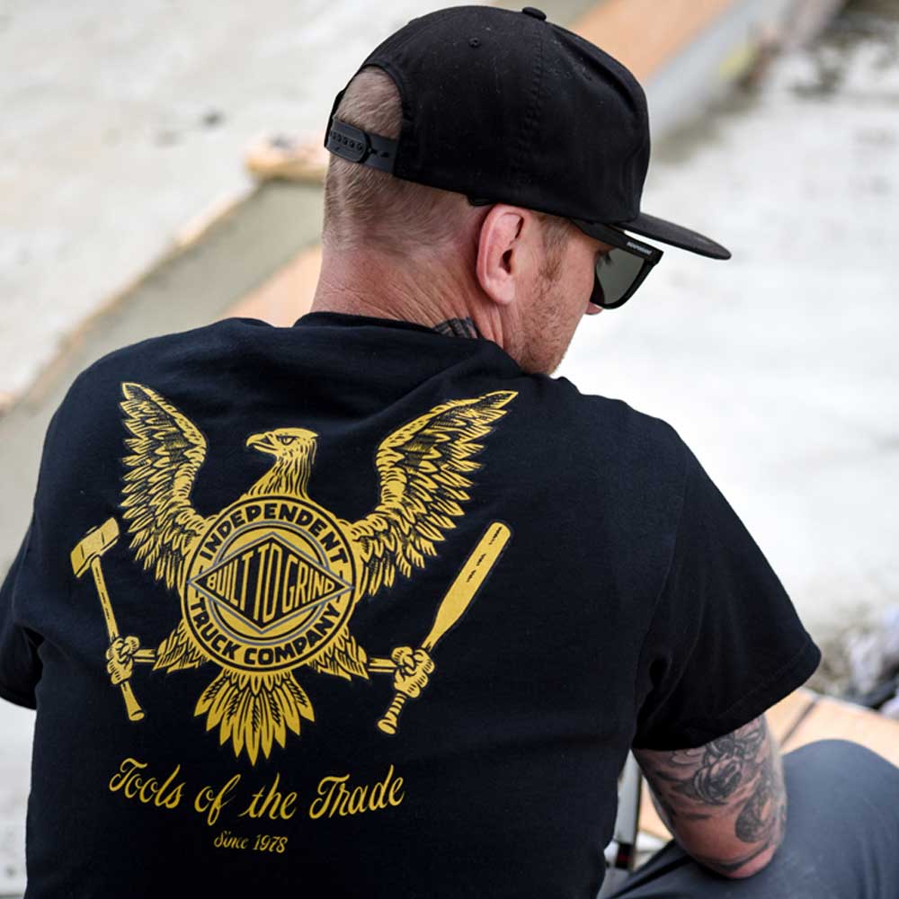 A man in a black cap and sunglasses, facing away, wears the Tools of the Trade S/S Heavyweight T-Shirt by Independent. The black tee features a bold yellow eagle with tools and “Independent Truck Company” on the back.