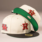 Two cream-colored The Cougar 2.0 Houston Astros New Era Hat 5950 Fitted caps are shown, both with black brims, green undersides, and red “H” logos on the front and side—one resting atop the other.