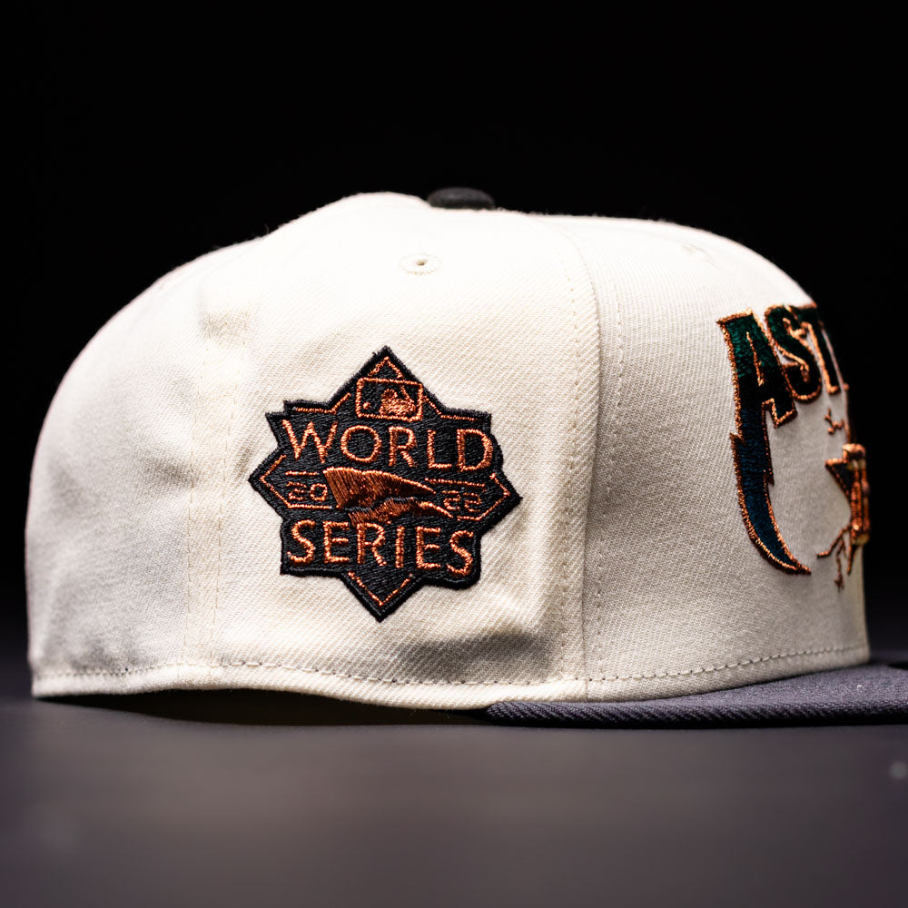 The Undaground 22WS Chrome Houston Astros New Era Hat 5950 Fitted is a white New Era baseball cap adorned with a black and orange World Series patch on the side and partial AST lettering on the front, honoring the Houston Astros. It is elegantly photographed against a dark background.