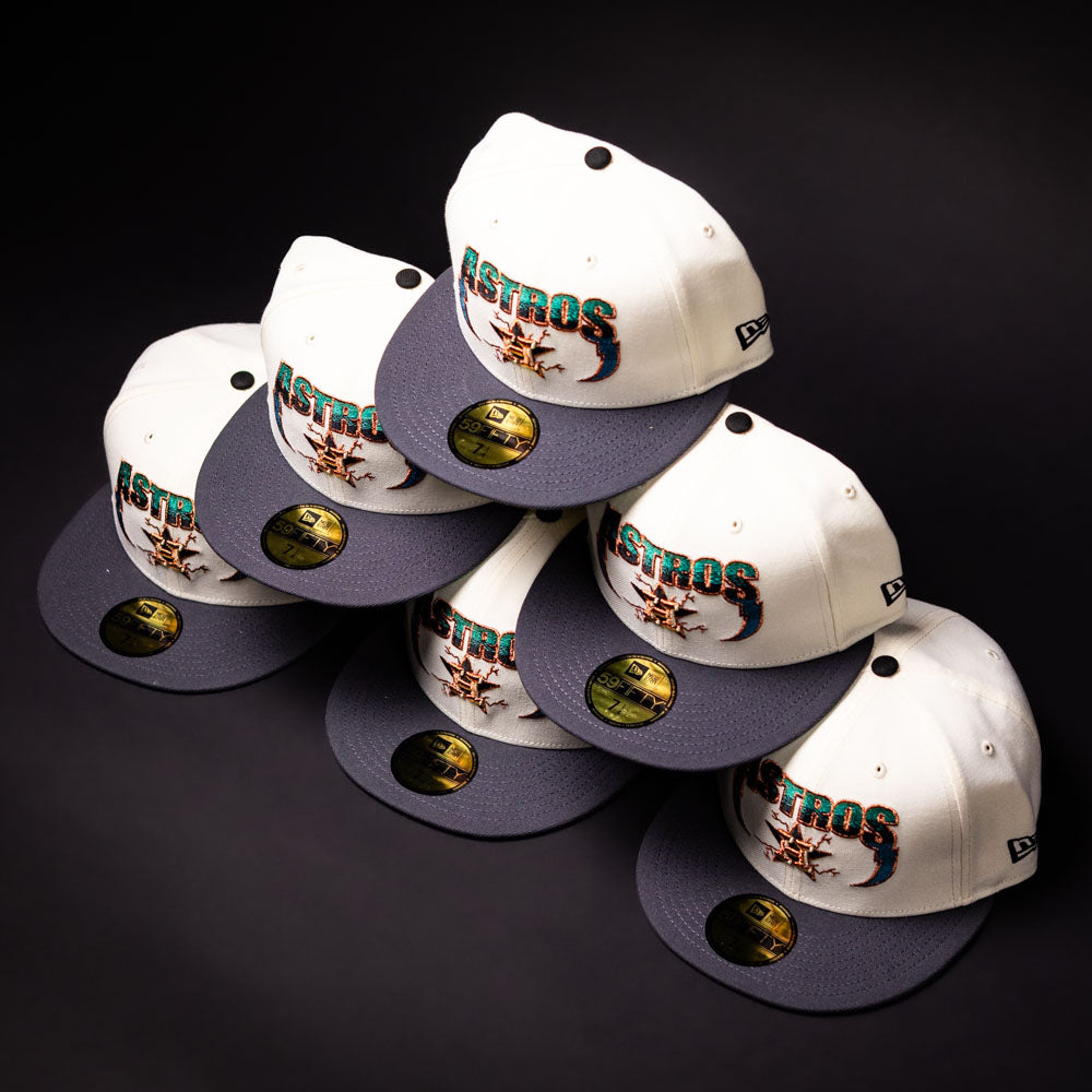 Six white and gray Undaground 22WS Chrome Houston Astros New Era Hat 5950 Fitted caps, each featuring a gold New Era sticker on the visor, are neatly stacked in a pyramid shape on a dark surface—showcasing the distinctive style of this exclusive collection.