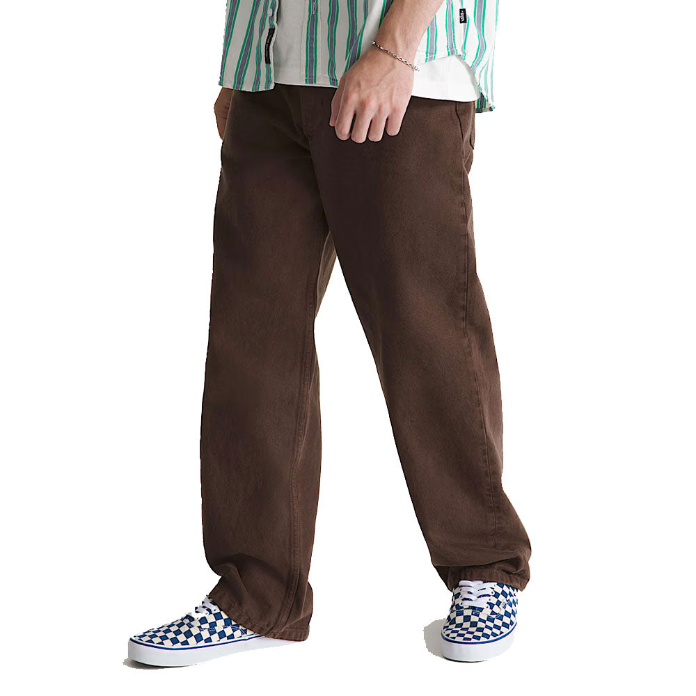 A person wearing Vans Loose Denim Dark Brown Check 5 Pants, a green and white striped shirt, and blue-and-white checkered slip-on shoes, standing against a white background.