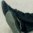 A close-up shows someone in dark pants wearing Vans Skate Half Cab Walnut Gum VN0A5FCD77J1 suede skateboard shoes with textured soles and PopCush impact protection, sitting cross-legged on a light, rough surface.