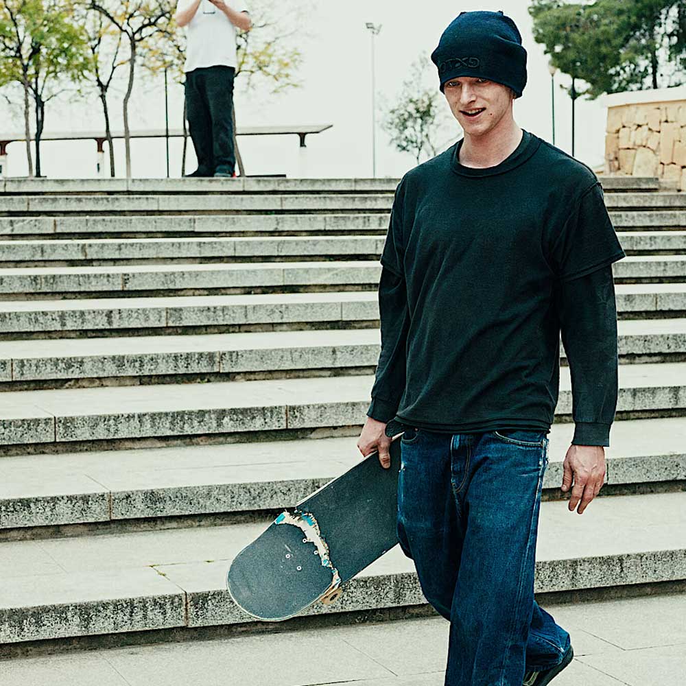 A young man in a black beanie, dark tee, jeans, and Vans Skate Half Cab Walnut Gum VN0A5FCD77J1 suede skateboard shoes walks down stone steps with a skateboard. Another person stands at the top; trees and a stone wall are seen behind.
