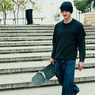 A young man in a black beanie, dark tee, jeans, and Vans Skate Half Cab Walnut Gum VN0A5FCD77J1 suede skateboard shoes walks down stone steps with a skateboard. Another person stands at the top; trees and a stone wall are seen behind.