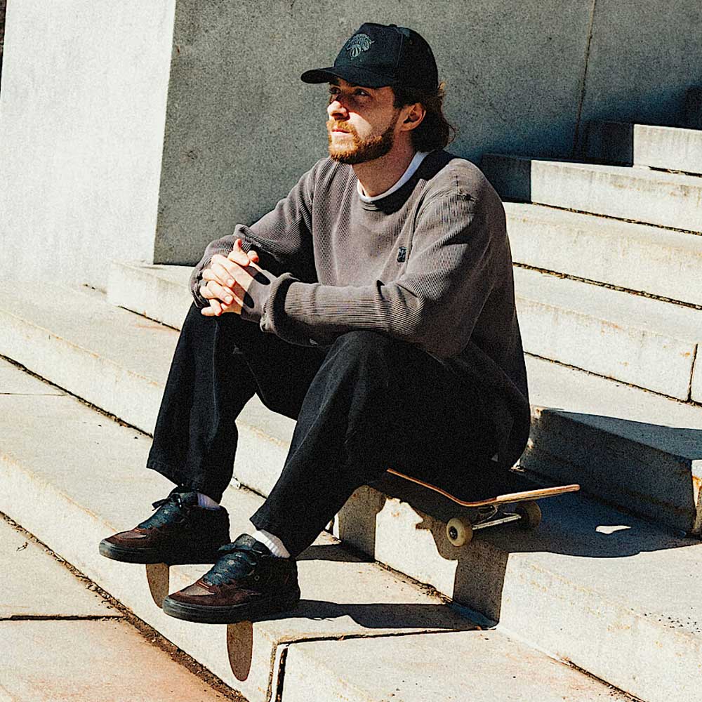 A bearded man in a black cap, gray sweater, and black pants sits thoughtfully on outdoor steps with a skateboard, wearing Vans Skate Half Cab Walnut Gum VN0A5FCD77J1 suede skateboard shoes for PopCush impact protection.