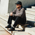 A bearded man in a black cap, gray sweater, and black pants sits thoughtfully on outdoor steps with a skateboard, wearing Vans Skate Half Cab Walnut Gum VN0A5FCD77J1 suede skateboard shoes for PopCush impact protection.