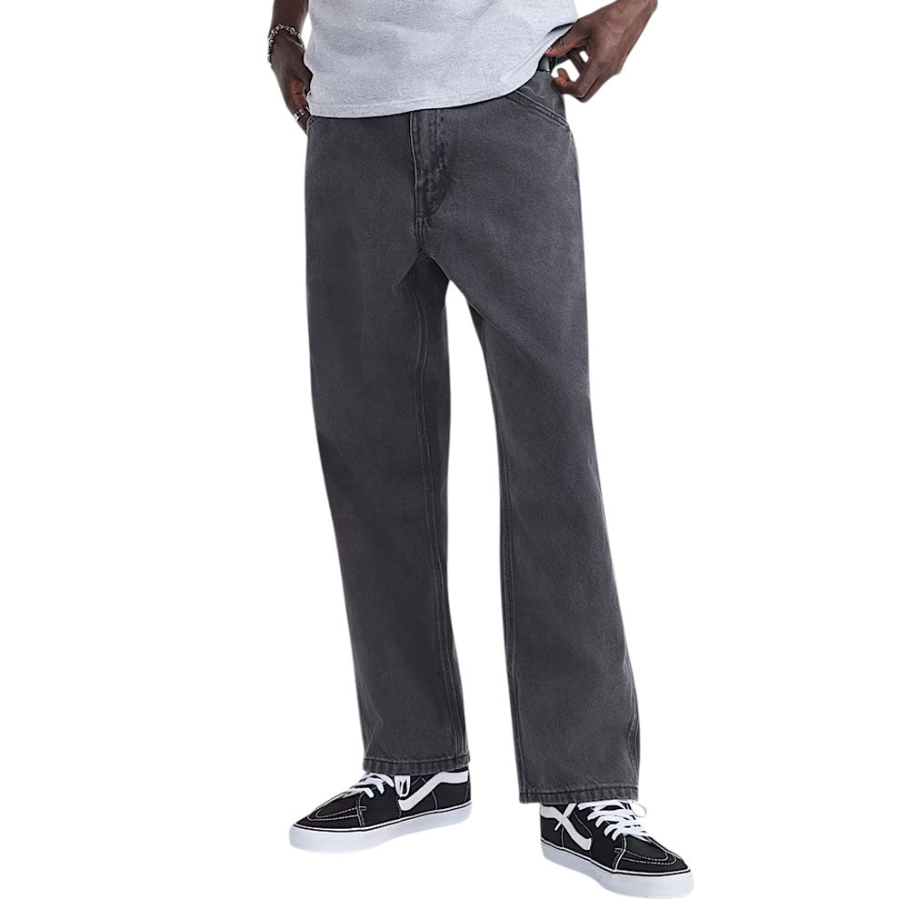 Someone wearing Vans Loose Denim Black VN000PH6BLK Skate Check 5 Pants, a light gray t-shirt, and black and white sneakers stands with one hand in their pocket against a plain background.