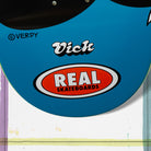 Close-up of the Verdy Vick 8.5 Real Skateboard Deck Skateshop Day, showing "Vick" and "REAL SKATEBOARDS" in bold inside an orange and white oval, plus a ©VERDY mark nearby.