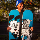 A smiling person in a blue beanie holds two skateboard decks, including the Verdy Vick 8.5 Real Skateboard Deck Skateshop Day, outdoors with trees in the background.