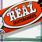 The Verdy Oval 8.38 Real Skateboard Deck Skateshop Day features a bold, cartoon-style REAL logo in white and black on a vivid orange background, accented with playful, eye-like details on the letters.
