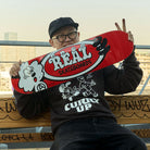 A smiling person in glasses, a black cap, and a dark sweatshirt sits on a bench holding the Verdy Oval 8.06 Real Skateboard Deck Skateshop Day—a limited edition release featuring bold red color and cartoon graphics.