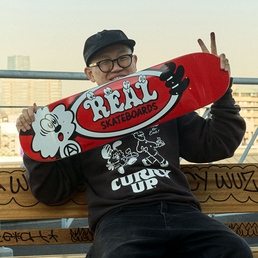 A smiling person in glasses and a black cap sits on a bench, holding the Verdy Oval 8.38 Real Skateboard Deck Skateshop Day with cartoon graphics. Wearing a dark “Curry Up” hoodie, they flash a peace sign.