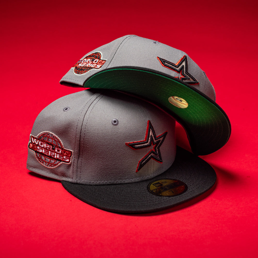 Two Days B4 WS05 Misty Black Houston Astros New Era 5950 Fitted hats are displayed on a vibrant red background. Both fitted caps feature a bold star logo on the front and a World Series patch on the side. One hat is stacked atop the other, highlighting the signature Misty Black brim and its green underside.