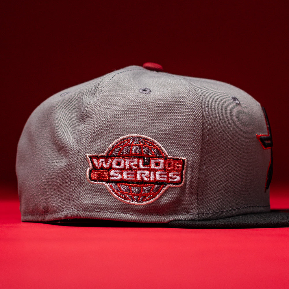 A Days B4 WS05 Misty Black Houston Astros New Era Hat 5950 Fitted rests on a red surface, featuring a gray crown and Misty Black brim. The side is adorned with a red and white World Series embroidered patch showcasing a globe design, emphasizing the hat’s authentic New Era craftsmanship. A solid red background completes the scene.