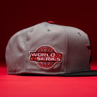 A Days B4 WS05 Misty Black Houston Astros New Era Hat 5950 Fitted rests on a red surface, featuring a gray crown and Misty Black brim. The side is adorned with a red and white World Series embroidered patch showcasing a globe design, emphasizing the hat’s authentic New Era craftsmanship. A solid red background completes the scene.