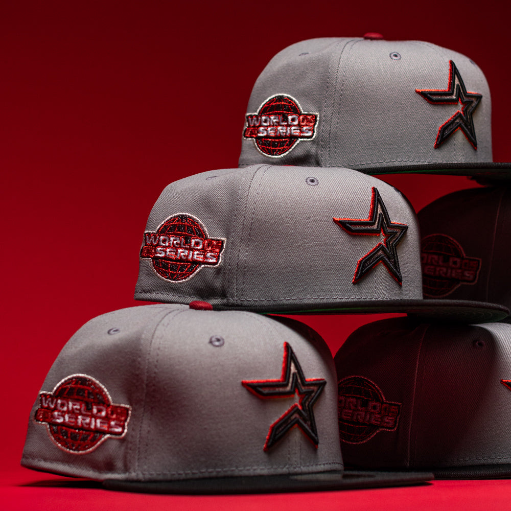 Gray New Era baseball caps featuring World Series logos and star patches are stacked in a pyramid shape against a red background, including the special edition Days B4 WS05 Misty Black Houston Astros New Era Hat 5950 Fitted.