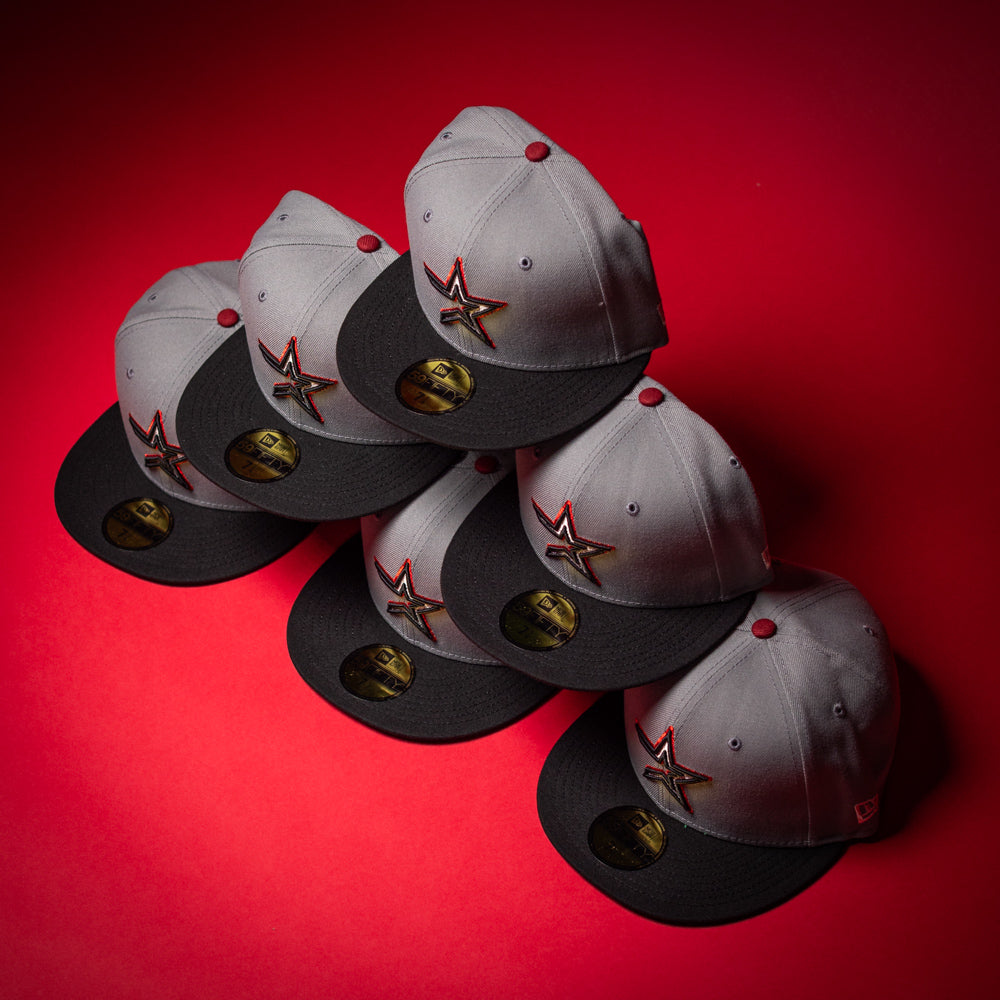 Six Days B4 WS05 Misty Black Houston Astros New Era Hat 5950 Fitted caps, featuring gray and black hues with star logos and gold stickers, are neatly stacked in a pyramid shape on a vibrant red background.