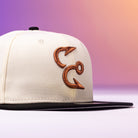 The Warren 2.0 Hooks Chrome Black New Era Hat 5950 Fitted features a cream-colored crown with a black visor, showcasing two embroidered fish hooks forming a stylized logo on the front. The smooth gradient background in purple and orange tones complements the bold Chrome Black accents.