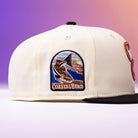 The Warren 2.0 Hooks Chrome Black New Era Hat 5950 Fitted features an embroidered patch of a fisherman holding a large fish by the coast, with “Coastal Bend” stitched below the image. The cream and black fitted cap stands out with a background blending purple, pink, and orange hues for a bold look.