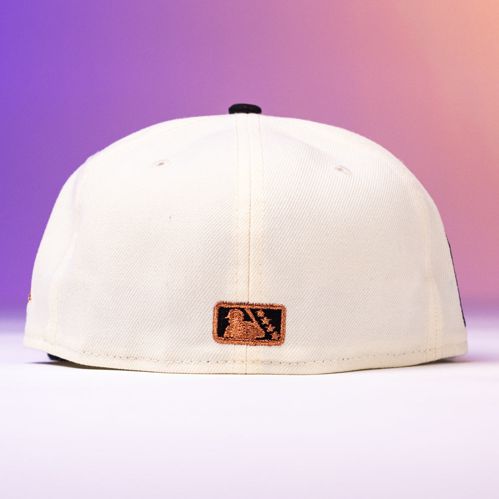 The Warren 2.0 Hooks Chrome Black New Era Hat 5950 Fitted is shown from the back, featuring a small brown patch with an embossed design. The modern cap stands out against a soft gradient background of purple, pink, and orange hues.