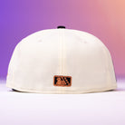The Warren 2.0 Hooks Chrome Black New Era Hat 5950 Fitted is shown from the back, featuring a small brown patch with an embossed design. The modern cap stands out against a soft gradient background of purple, pink, and orange hues.