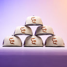 Six Warren 2.0 Hooks Chrome Black New Era Hat 5950 Fitted caps are stacked in a pyramid formation, their chrome black finish and signature style standing out against a vibrant purple and orange gradient background, showcasing the classic New Era fitted design.
