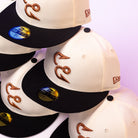Six cream and black Warren 2.0 Hooks Chrome Black New Era Hat 5950 Fitted caps featuring brown embroidery are stacked diagonally. The gold sticker on the brim displays 59FIFTY, 7 1/4, highlighting the signature Chrome Black fitted style. The background is light purple.