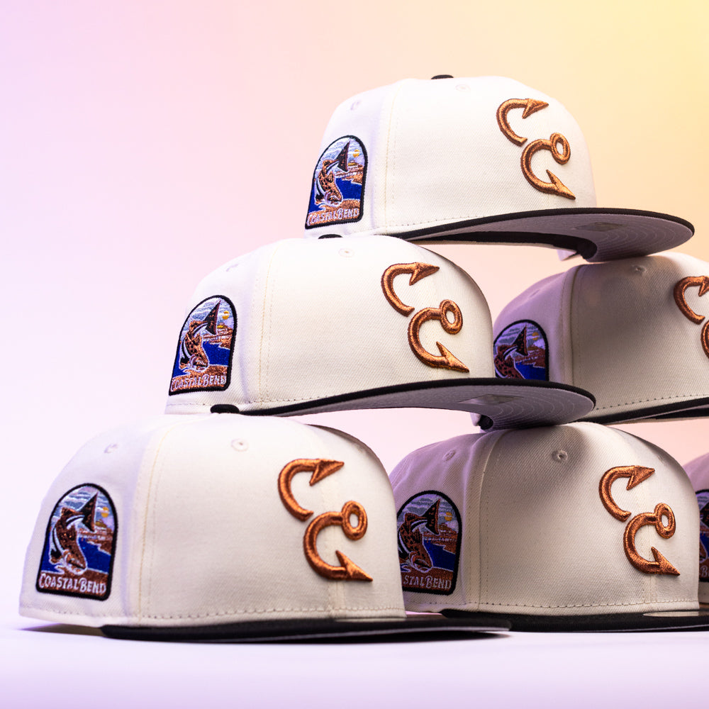 Six Warren 2.0 Hooks Chrome Black New Era Hat 5950 Fitted caps, featuring white crowns with black bills, are stacked in a pyramid. Each cap showcases embroidered fishing hooks on the side and a Coastal Bend patch with a fish and wave design on the front, all arranged against a softly lit, pastel-hued background for an eye-catching display of Chrome Black style.
