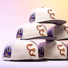 Six Warren 2.0 Hooks Chrome Black New Era Hat 5950 Fitted caps, featuring white crowns with black bills, are stacked in a pyramid. Each cap showcases embroidered fishing hooks on the side and a Coastal Bend patch with a fish and wave design on the front, all arranged against a softly lit, pastel-hued background for an eye-catching display of Chrome Black style.
