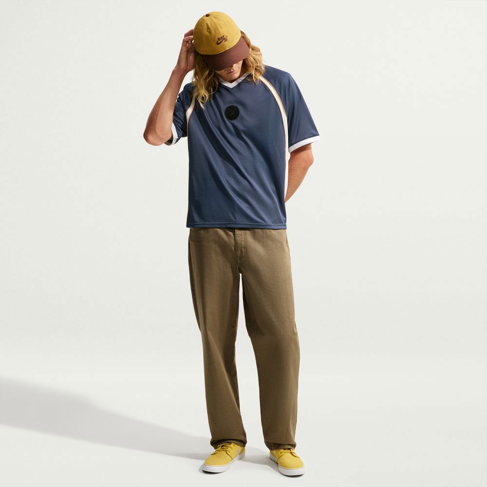 A person with long hair wears a yellow cap, navy polo shirt, khaki pants, and yellow shoes—standing against a plain white background, looking down with one hand on the cap. The outfit is paired with the Yuto 1 Jersey Navy (IM5465-437), a top from Nike SB Skateboarding featuring sweat-wicking fabric for added comfort.