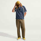 A person with long hair wears a yellow cap, navy polo shirt, khaki pants, and yellow shoes—standing against a plain white background, looking down with one hand on the cap. The outfit is paired with the Yuto 1 Jersey Navy (IM5465-437), a top from Nike SB Skateboarding featuring sweat-wicking fabric for added comfort.