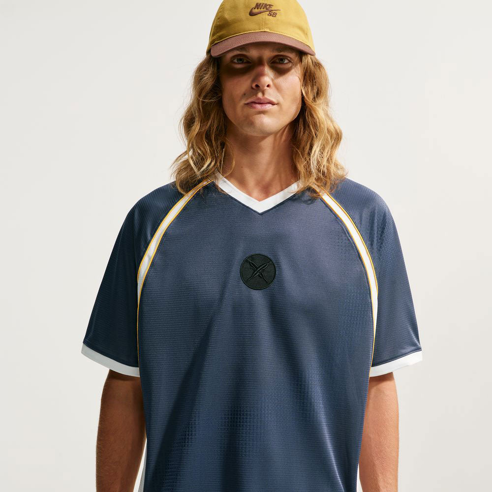 A person with long blond hair wears a yellow cap and the Nike SB Yuto 1 Jersey Navy (IM5465-437), a dark blue V-neck skateboarding top featuring yellow and white trim and sweat-wicking properties, while standing against a plain light background.
