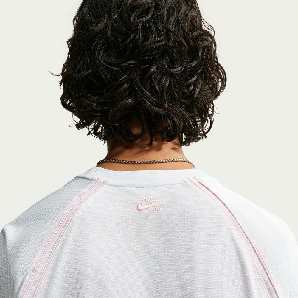 A person with curly dark hair, seen from behind, wears the Nike SB Yuto 1 Jersey White (IM5465-100), featuring pink stitching and a silver chain necklace. The back of this skateboarding top displays a small pink Nike SB logo near the neckline, while its sweat-wicking fabric ensures comfort both on and off the board.