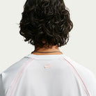 A person with curly dark hair, seen from behind, wears the Nike SB Yuto 1 Jersey White (IM5465-100), featuring pink stitching and a silver chain necklace. The back of this skateboarding top displays a small pink Nike SB logo near the neckline, while its sweat-wicking fabric ensures comfort both on and off the board.