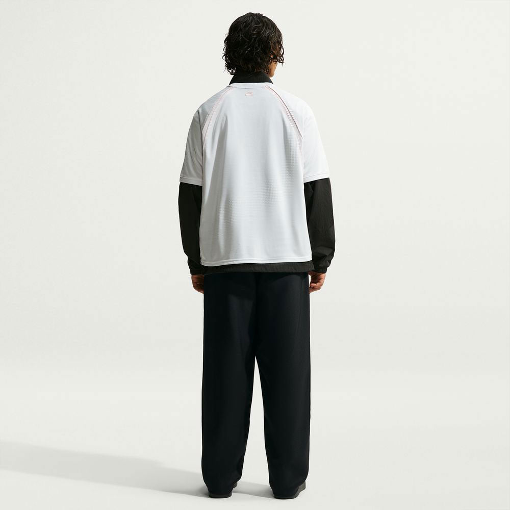 A person stands facing away against a plain light background, wearing the Nike SB Yuto 1 Jersey White IM5465-100 top over a long-sleeve black shirt and loose black pants, with short dark hair.