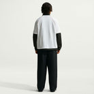 A person stands facing away against a plain light background, wearing the Nike SB Yuto 1 Jersey White IM5465-100 top over a long-sleeve black shirt and loose black pants, with short dark hair.