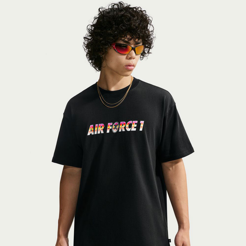 Person with curly hair wearing red sunglasses, gold chains, and a black Yuto 1 Tee (IM4432-010) from Nike SB Skateboarding—with AIR FORCE 1 in colorful letters—styled similarly to a Nike SB Jacquard Skate Jersey, standing against a plain light background.