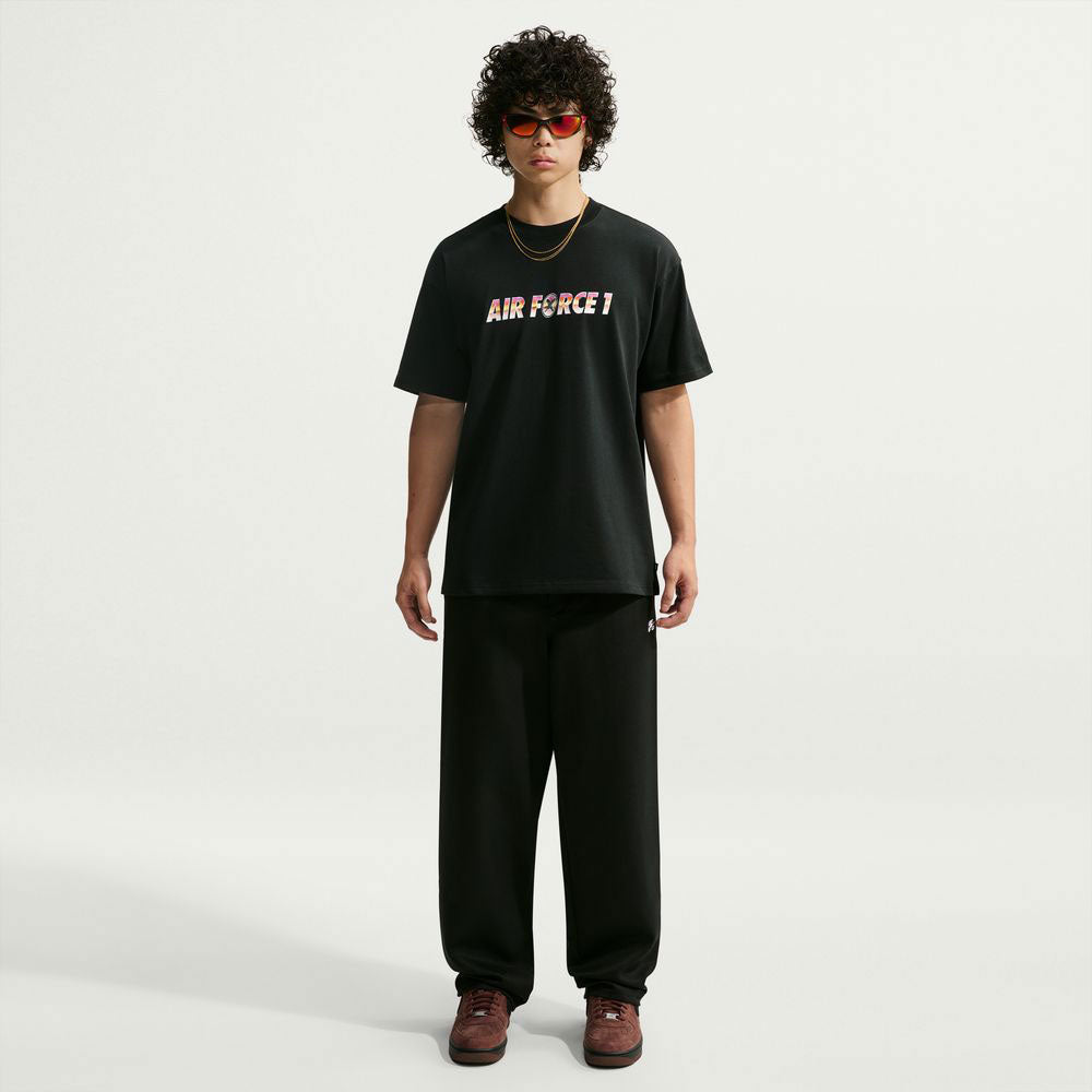 A person with curly hair wearing red sunglasses, a black Yuto 1 Tee Black IM4432-010 T-Shirt from Nike SB Skateboarding known for its sweat-wicking fabric, loose black pants, brown shoes, and gold chains stands against a plain white background.