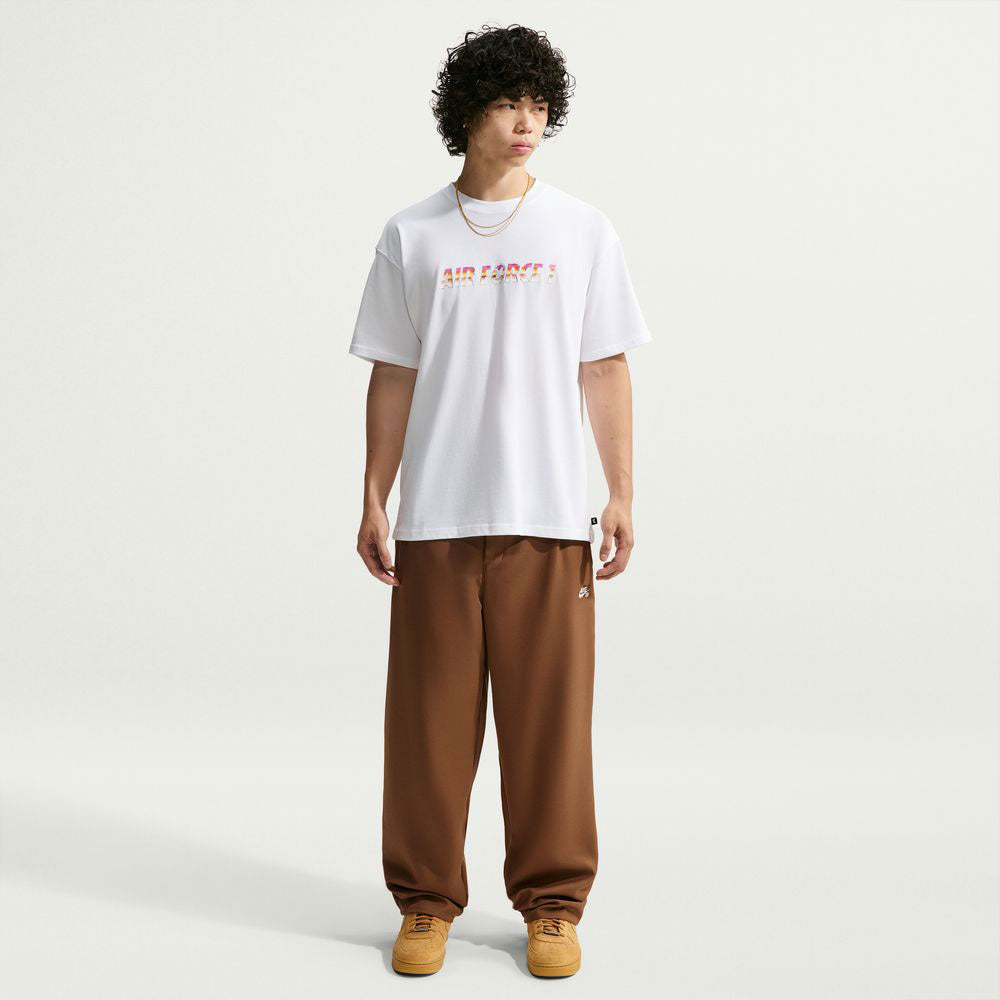 A person with curly hair wears a white Yuto 1 Tee White IM4432-100 T-Shirt from Nike SB Skateboarding, loose brown pants, tan sneakers, and a gold chain, standing against a plain light background.