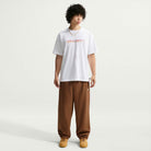 A person with curly hair wears a white Yuto 1 Tee White IM4432-100 T-Shirt from Nike SB Skateboarding, loose brown pants, tan sneakers, and a gold chain, standing against a plain light background.