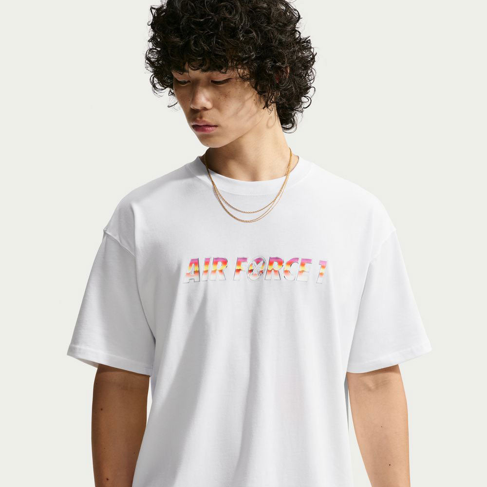 A young person wearing the Yuto 1 Tee White IM4432-100 T-Shirt from Nike SB Skateboarding, featuring “AIR FORCE 1” in colorful lettering and two gold chains, looks down with curly hair against a plain light background—capturing the effortless cool and skate-inspired vibe of a sweat-wicking Nike SB top.