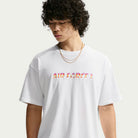 A young person wearing the Yuto 1 Tee White IM4432-100 T-Shirt from Nike SB Skateboarding, featuring “AIR FORCE 1” in colorful lettering and two gold chains, looks down with curly hair against a plain light background—capturing the effortless cool and skate-inspired vibe of a sweat-wicking Nike SB top.