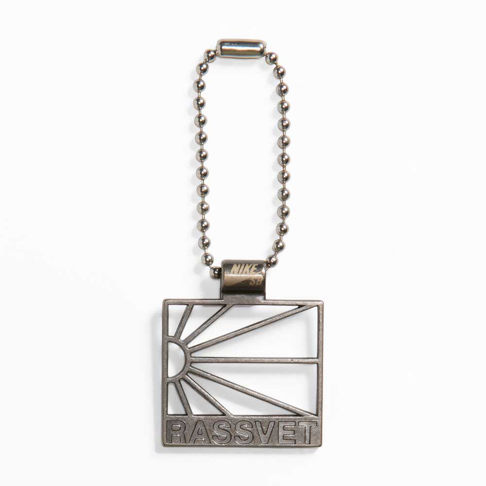 A metallic keychain with a square pendant featuring a stylized rising sun and "RASSVET" engraving, inspired by the Zoom Tennis Classic x Rassvet Blue Void Sail Nike SB Leather Skateboarding Shoes, attached to a beaded chain.