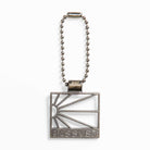 A metallic keychain with a square pendant featuring a stylized rising sun and "RASSVET" engraving, inspired by the Zoom Tennis Classic x Rassvet Blue Void Sail Nike SB Leather Skateboarding Shoes, attached to a beaded chain.