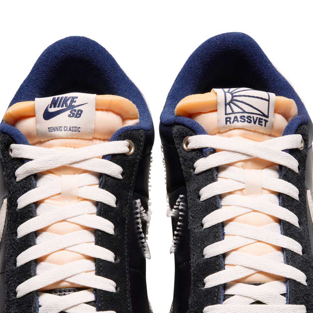 Close-up of Zoom Tennis Classic x Rassvet IO9846400 Blue Void Sail Nike SB leather skate shoes with white laces. The left tongue shows the Nike SB logo, while the right features the Rassvet sun logo on a white patch.