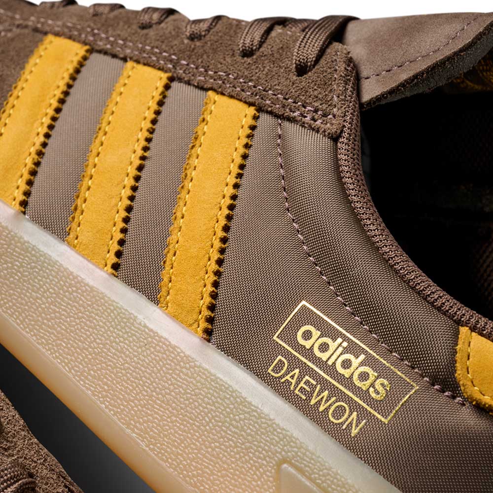 Close-up of the Daewon Song Glenburn Brown Yellow KZ8777 Adidas Skateboarding Shoe, featuring gold stripes and “adidas DAEWON” text in gold above the sole. The image captures only part of the shoe, emphasizing its classic adidas trainers design and understated skate style.
