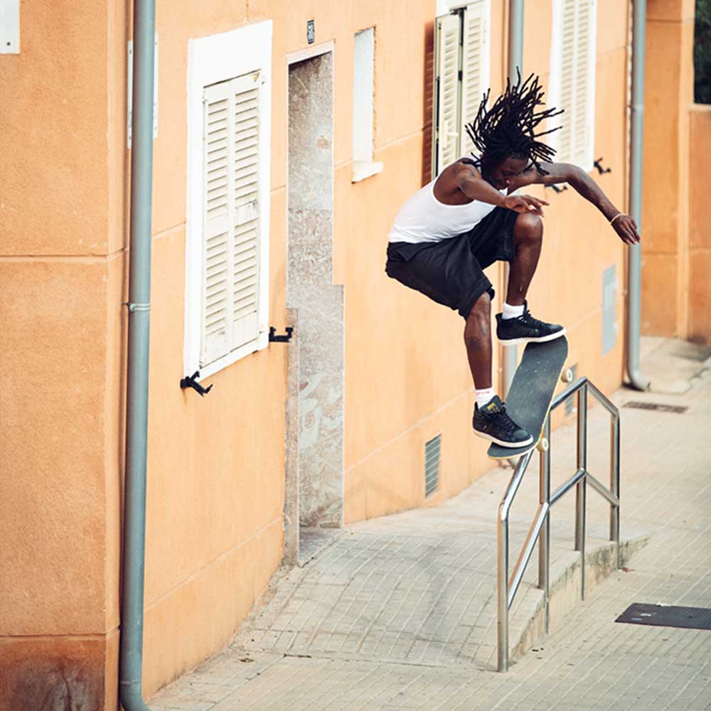 A skateboarder with long dreadlocks jumps over a handrail outside an orange building, wearing a white tank top, black shorts, and adidas Superskate x Kader Shoes JQ4413 in Core Black/Off White suede and leather.